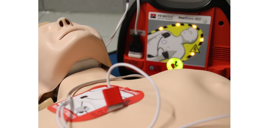 Man sieht eine Übungspuppe und einen Defibrillator.