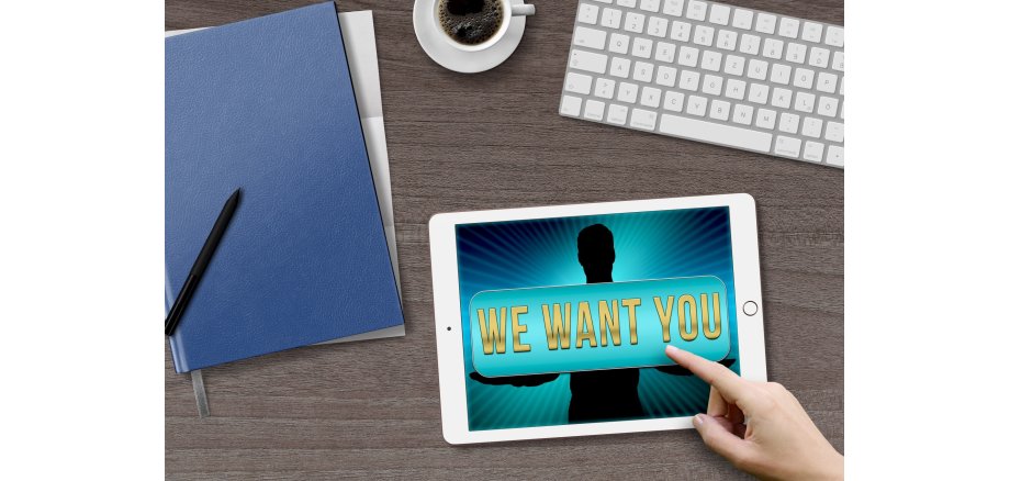 Auf dem Bild sieht man einen Laptop und Schreibmaterial sowie ein Schriftzug auf dem steht "We want you"