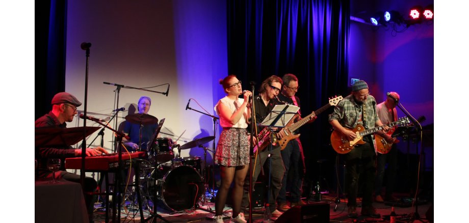 Man sieht eine Band die Instrumente spielt und eine Frau die singt.
