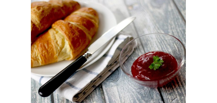 Auf dem Bild sieht man einen Teller mit Croissants, Besteck und ein Schälchen mit Erdbeermarmelade