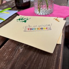 Man sieht ein Blatt Papier mit dem Wort "Tschüss" in bunten Farben sowie gezeichneten Figuren. 