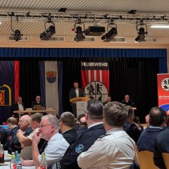 Man sieht mehrere Menschen auf der Bühne. Im Hintergrund hängt ein Banner von der Feuerwehr.