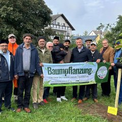 Auf dem Bild sieht man den Bürgermeister Dominik Stadler und eine Gruppe von Menschen die einen Banner mit der Aufschrift "Baumpflanzung" hochhalten.