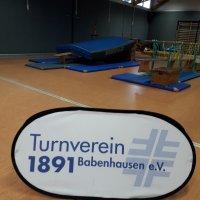 Man sieht das blaue Logo des Turnverein Babenhausen e.V. Im Hintergrund ist eine Sporthalle mit Matten.